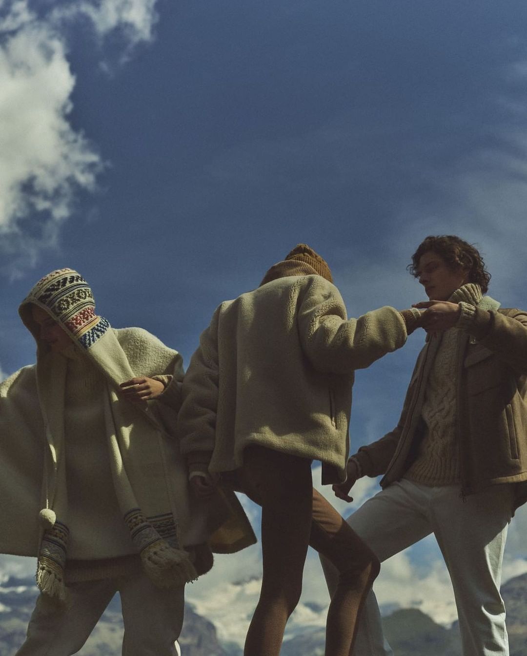 Three people in outdoor fashion shot with blue skies and mountains. Two on left wear patterned headwear, one on right wears brown jacket.