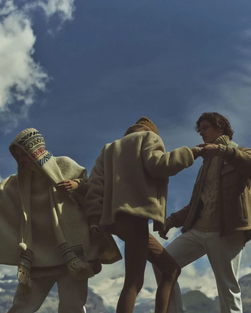 Three people in outdoor fashion shot with blue skies and mountains. Two on left wear patterned headwear, one on right wears brown jacket.