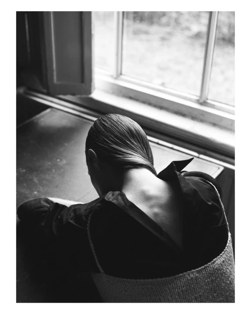 Back view of an adult female with dark hair slicked back, wearing a dark top and light-colored woven garment, sitting indoors near a window.