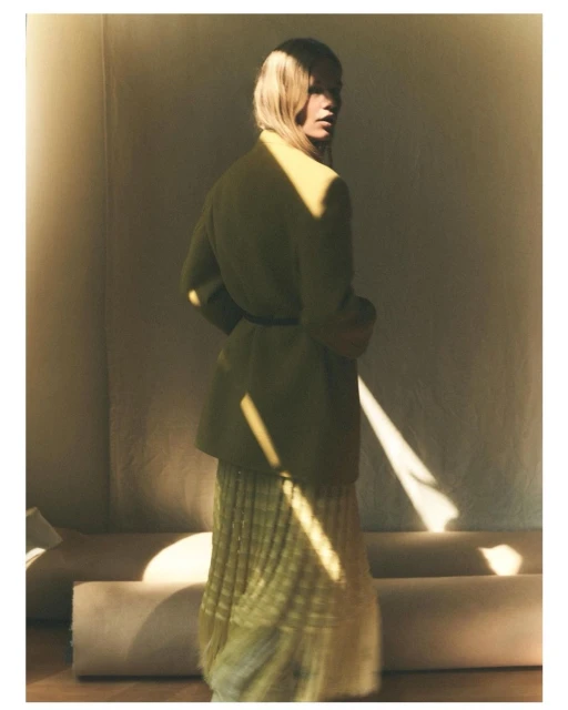 A woman in a yellow jacket and skirt, looking over her shoulder, stands in a naturally lit indoor setting with neutral tones.