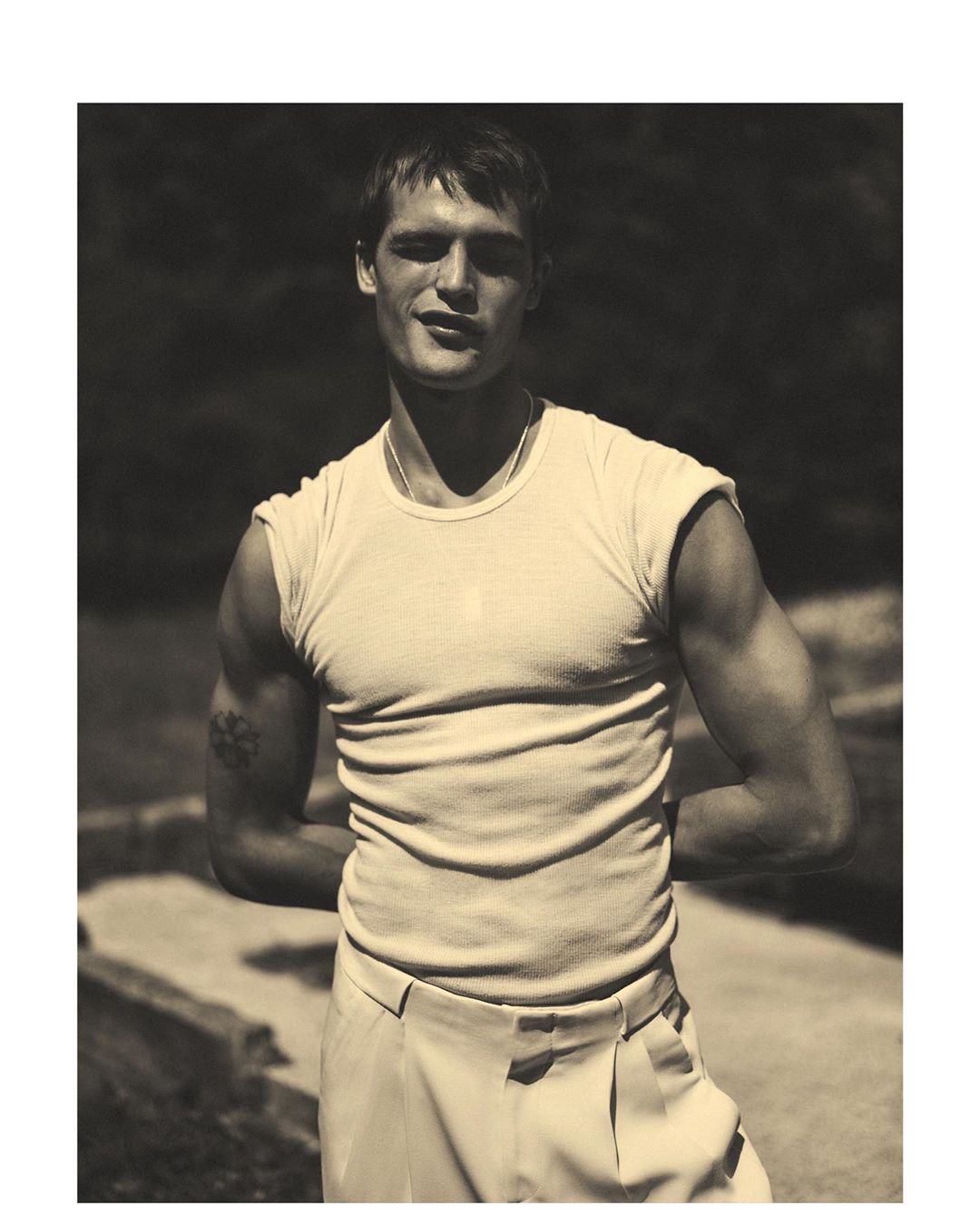 Man wearing a white ribbed t-shirt and light trousers posing outdoors with arms behind his back
