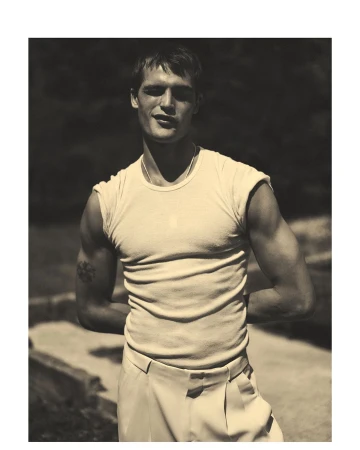 Man wearing a white ribbed t-shirt and light trousers posing outdoors with arms behind his back