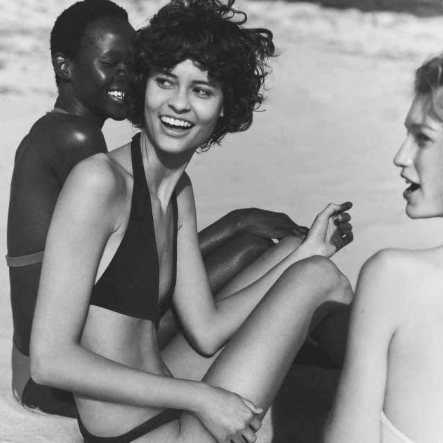 Three diverse adult women in swimsuits seated on a sandy beach, laughing and talking. Black and white photo with a shallow depth of field.