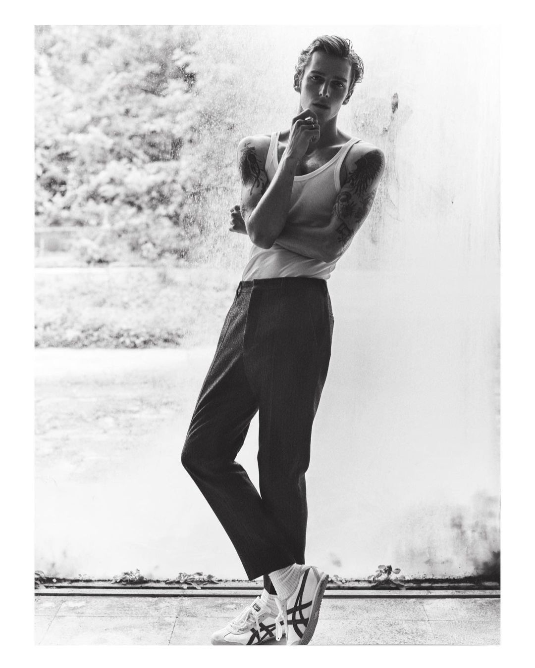 Monochrome portrait of a young man wearing a white tank top and dark trousers leaning against a textured wall