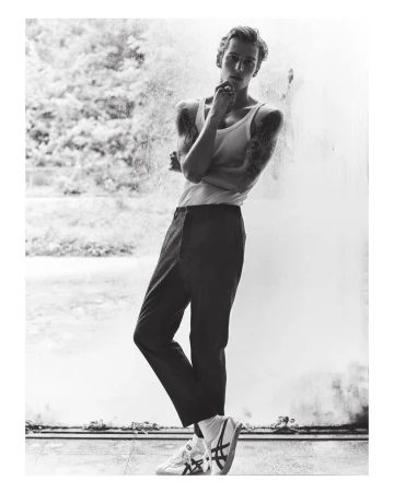 Monochrome portrait of a young man wearing a white tank top and dark trousers leaning against a textured wall