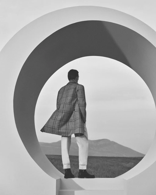 Monochrome shot of a man in a patterned coat and white pants viewed through a circular architectural element against a sky and mountains.