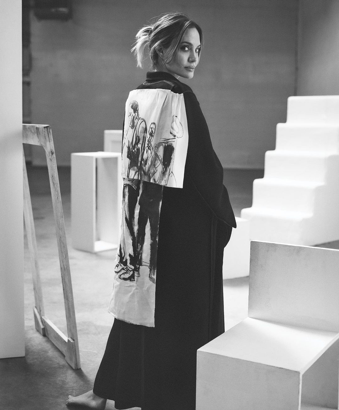 Adult woman in black dress and kimono with drawing, stands in a minimalist studio with white block furniture, looking at camera.