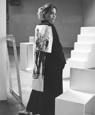 Adult woman in black dress and kimono with drawing, stands in a minimalist studio with white block furniture, looking at camera.