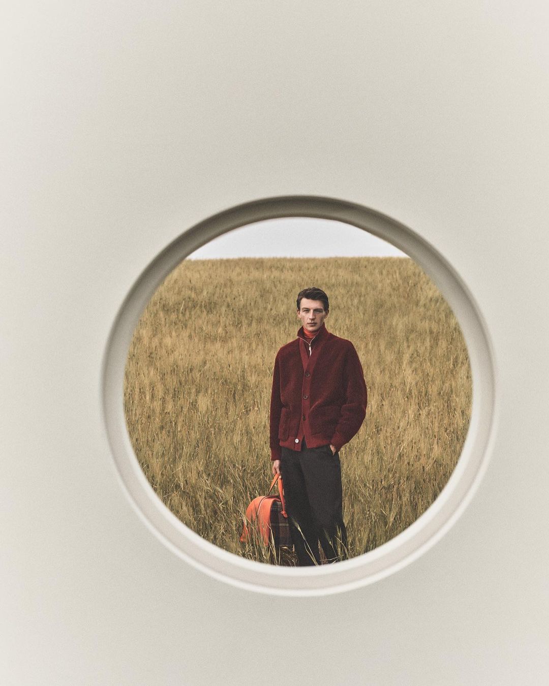 A man in a burgundy jacket is holding a tote bag, standing in a field of tall grass, seen through a white circular frame.