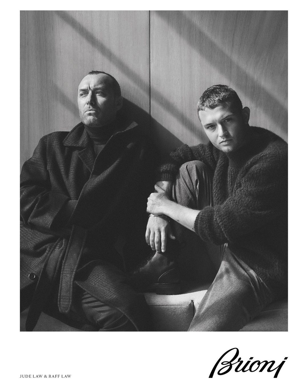 Black and white portrait of two adult males in coats seated in front of a panel wall with light and shadow, posing for a fashion ad.