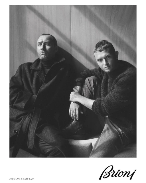 Black and white portrait of two adult males in coats seated in front of a panel wall with light and shadow, posing for a fashion ad.