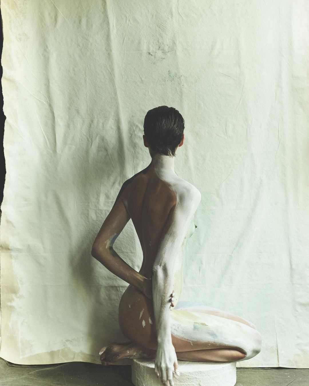 Back view of a nude adult female seated on a white platform, her body partially covered in white paint, against a draped white cloth backdrop.
