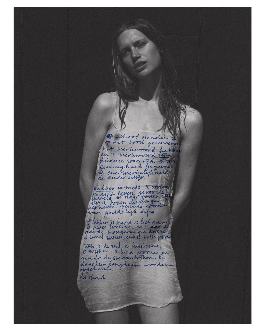 Artistic portrait of a young woman wearing a sheer dress with handwritten blue text, set against a dark wall. Fashion and style.