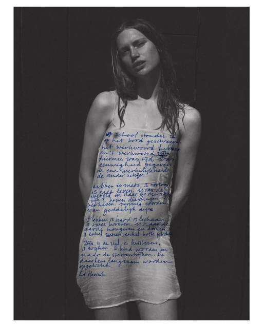 Artistic portrait of a young woman wearing a sheer dress with handwritten blue text, set against a dark wall. Fashion and style.