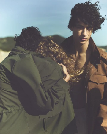 Portrait of a man with curly hair in a brown jacket standing near two people embracing outdoors by water and hills.