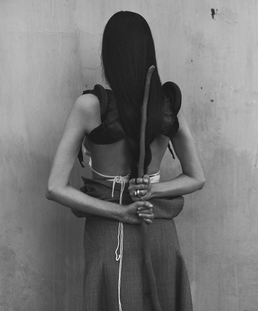 A woman stands with her back to the camera, wearing a skirt and carrying a log and black garment over her shoulders.