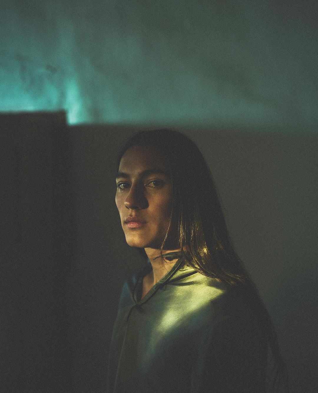 Portrait of an adult with long brown hair wearing a hooded shirt, illuminated by teal and yellow lighting indoors.