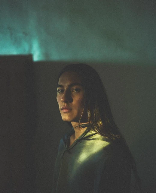 Portrait of an adult with long brown hair wearing a hooded shirt, illuminated by teal and yellow lighting indoors.
