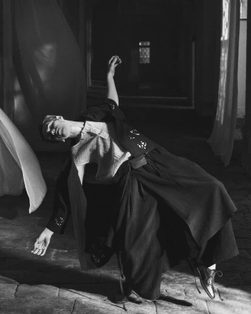 Black and white photo of a woman in flowing garments leaning to her right, one arm up, set indoors with window and curtain.