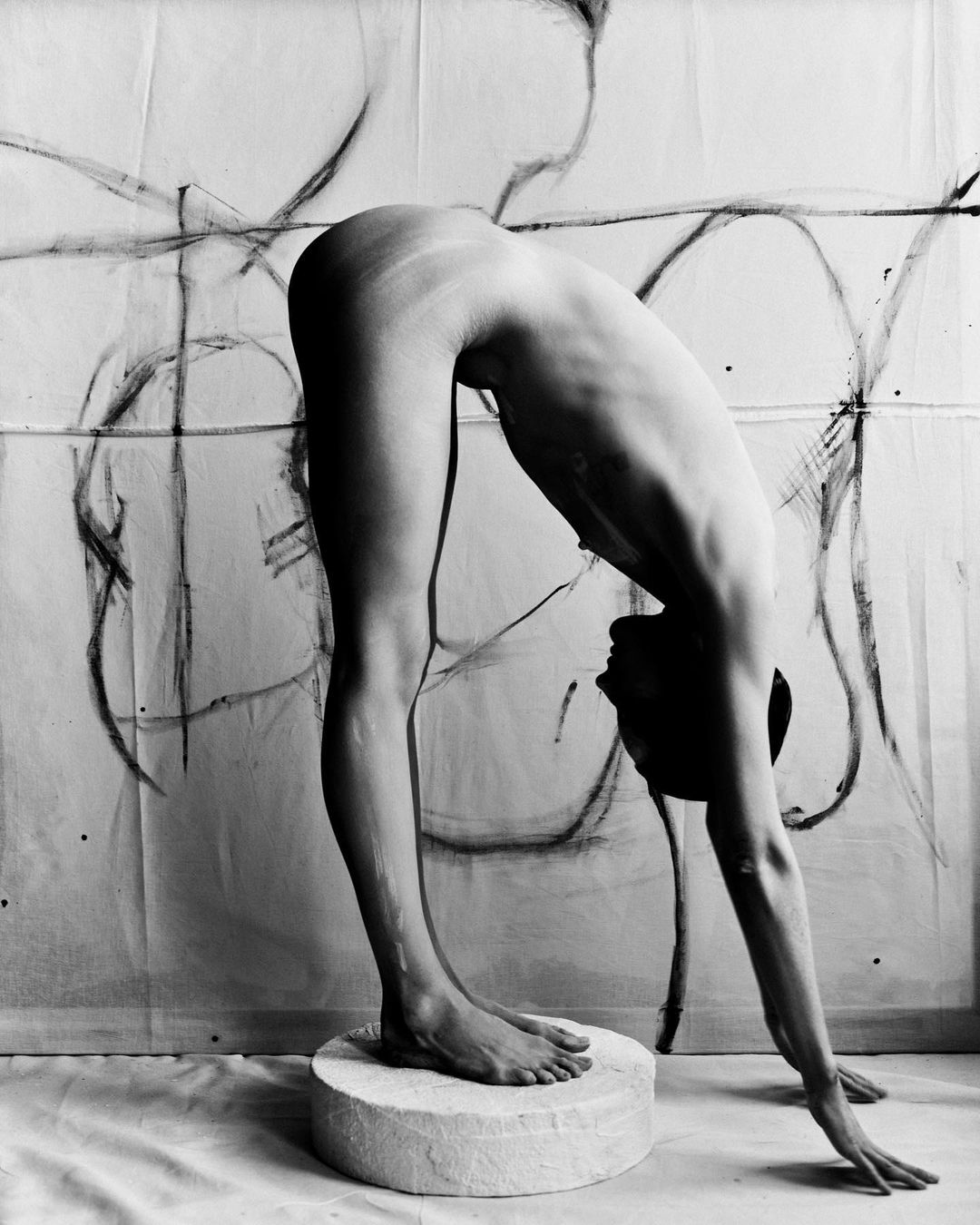 Monochrome image of a nude adult standing on a platform with arched back and hands reaching toward the floor, set against an abstract backdrop.