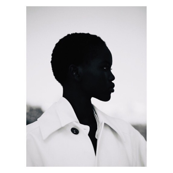 Monochromatic portrait of a dark-skinned woman in profile, wearing a white jacket against a neutral background, showcasing fashion and beauty.