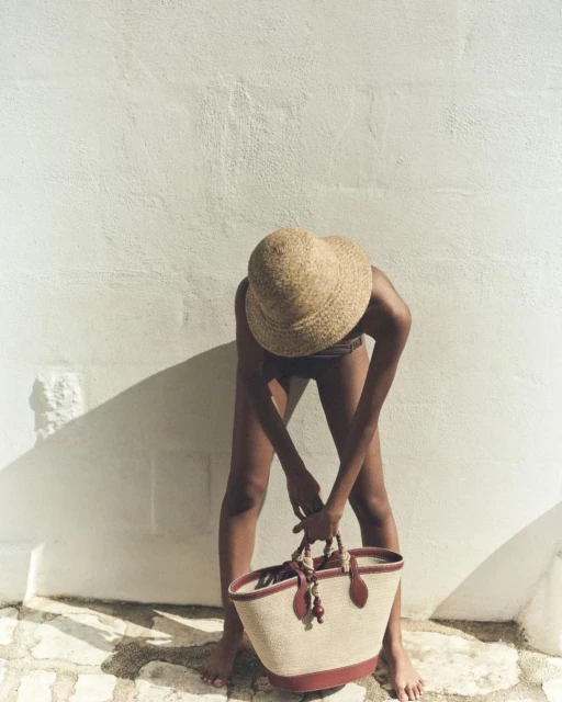 A woman in swimwear and a sunhat stands outdoors, holding a raffia tote bag against a white wall, on a sunny day casting soft shadows.