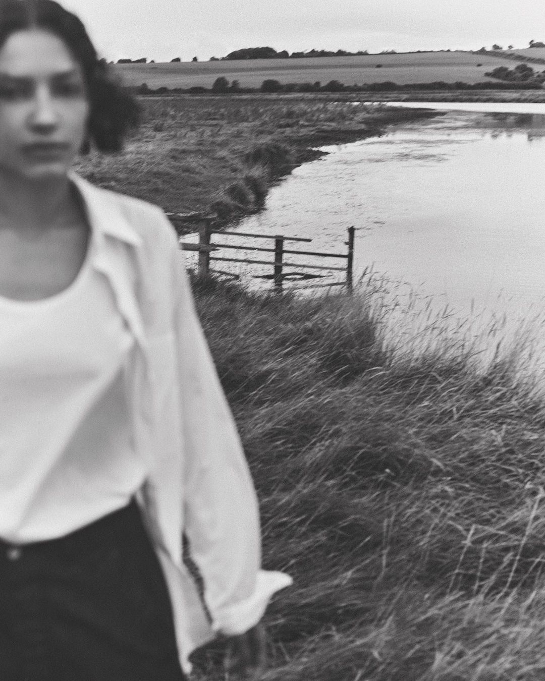 Black and white outdoor shot of a woman wearing an open white blouse, standing in tall grass near a body of water.