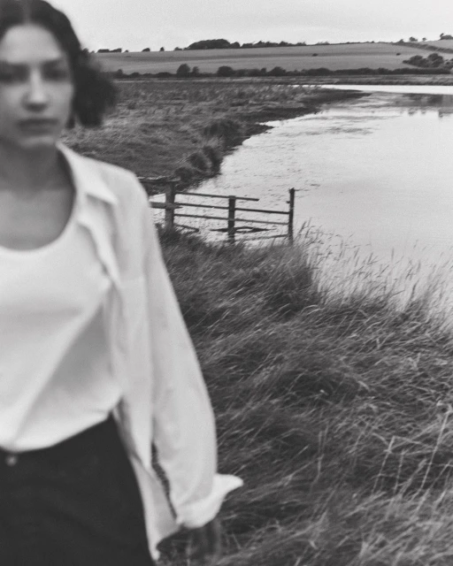 Black and white outdoor shot of a woman wearing an open white blouse, standing in tall grass near a body of water.