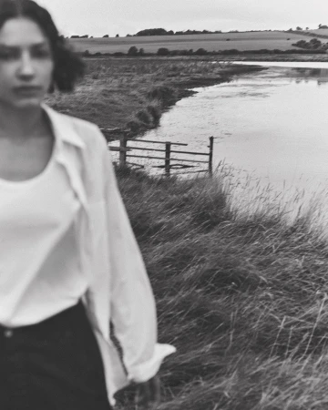 Black and white outdoor shot of a woman wearing an open white blouse, standing in tall grass near a body of water.
