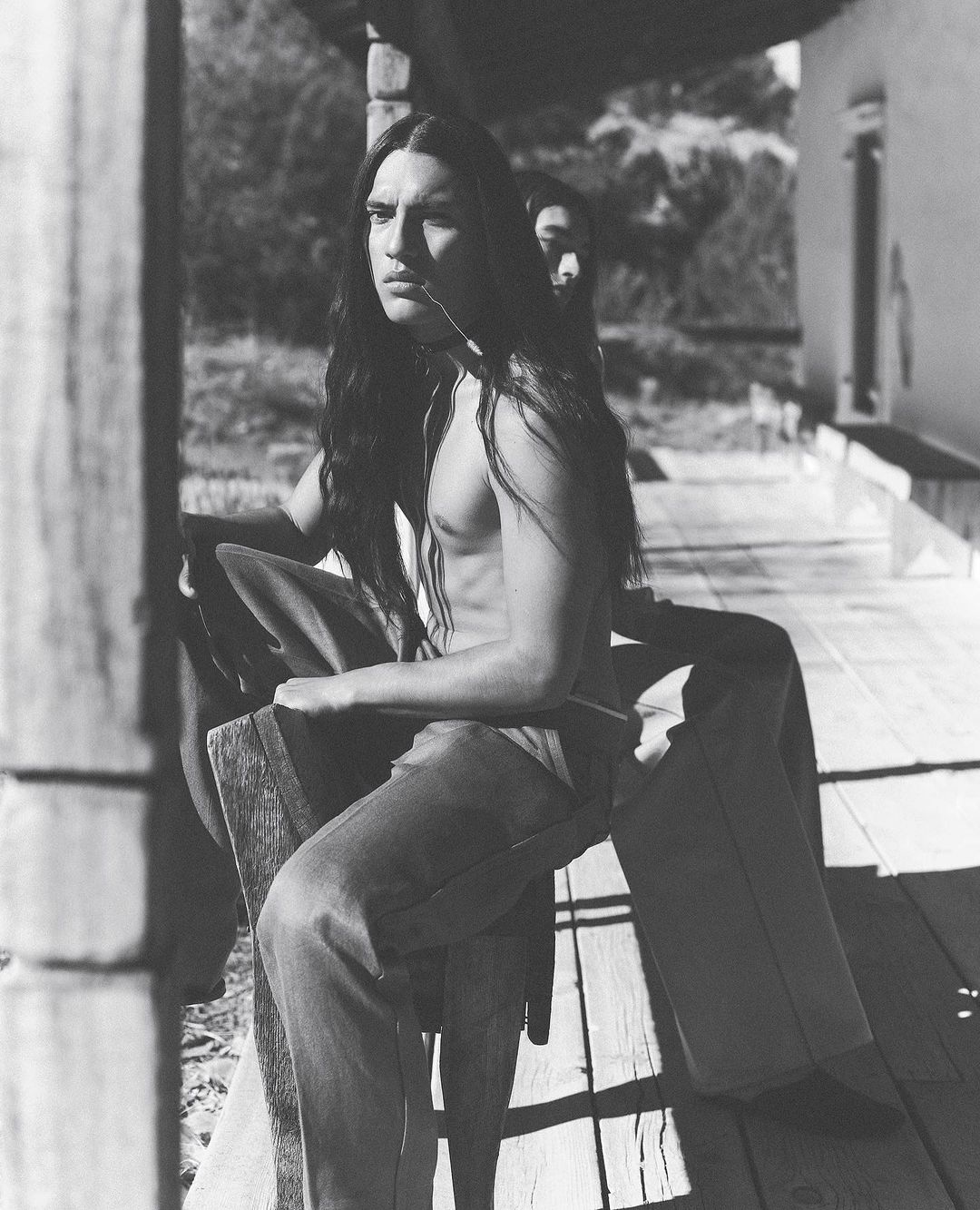 Monochrome image of a man with long hair sitting on a porch, contemplative expression, casual clothes, and another person in background.