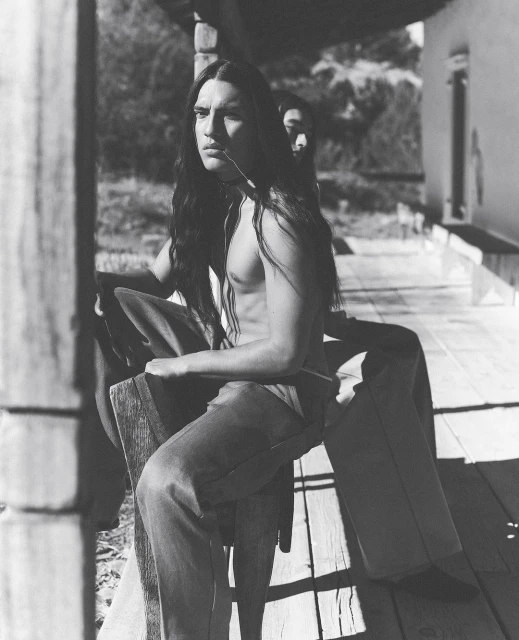 Monochrome image of a man with long hair sitting on a porch, contemplative expression, casual clothes, and another person in background.