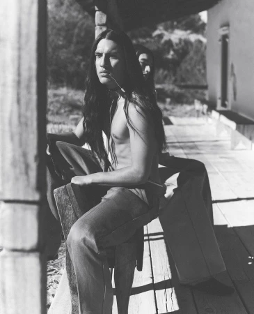 Monochrome image of a man with long hair sitting on a porch, contemplative expression, casual clothes, and another person in background.