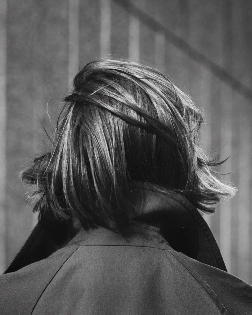 Monochrome portrait of an adult with short highlighted hair and a jacket, viewed from the rear against a blurred background.