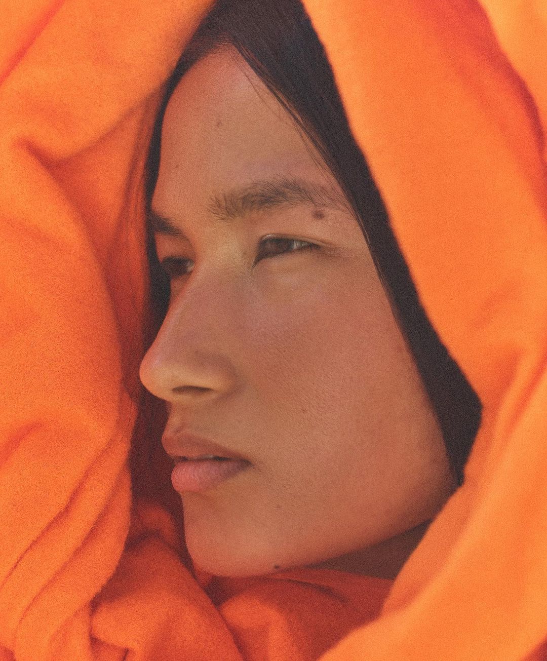 Close-up portrait of a person with dark hair and light skin wrapped in orange fabric, eyes looking away, illuminated by soft lighting.