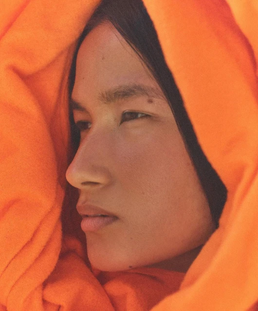 Close-up portrait of a person with dark hair and light skin wrapped in orange fabric, eyes looking away, illuminated by soft lighting.