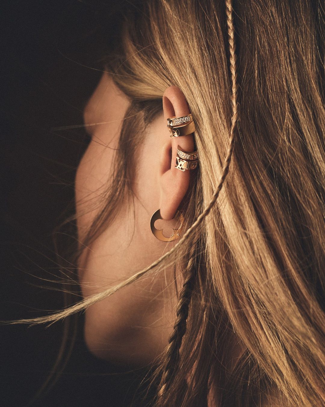 Close-up portrait of a woman wearing layered gold earrings and ear cuffs with long blonde hair and a small braid.