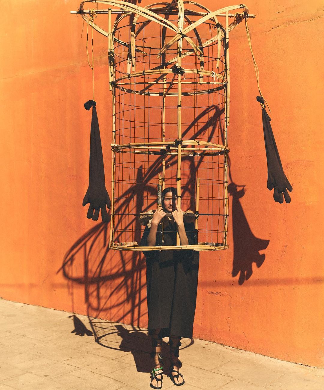 Person stands inside a large bamboo-framed figure in front of an orange wall, casting a shadow; the figure has glove-shaped appendages.