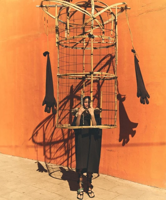 Person stands inside a large bamboo-framed figure in front of an orange wall, casting a shadow; the figure has glove-shaped appendages.