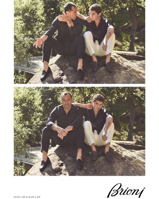 Two men seated on a rock in an outdoor setting, modeling menswear. One man wears a navy suit, the other wears white pants and a navy shirt.