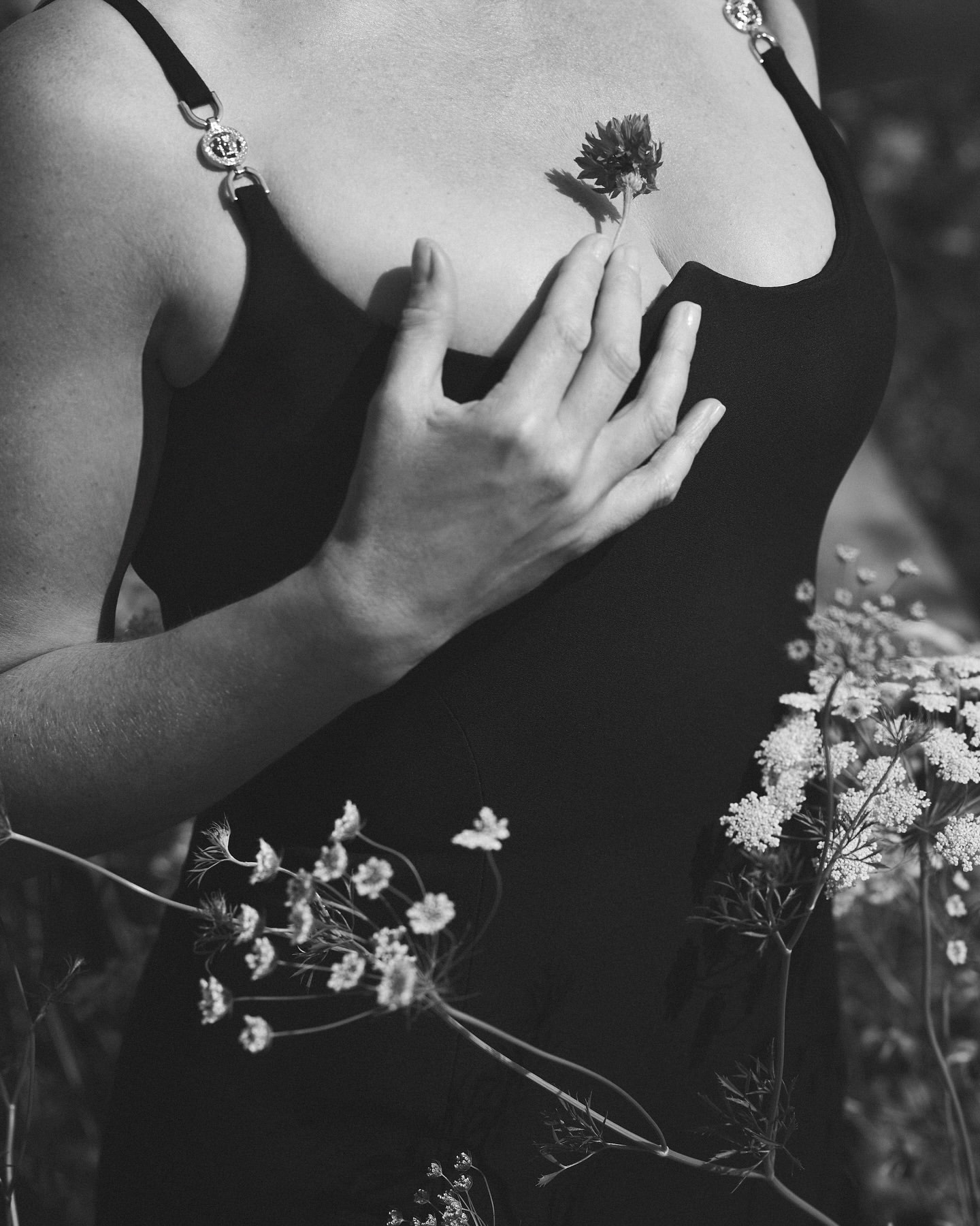Monochrome close-up of a woman in a black strap dress holding a small clover flower against her chest among wildflowers.