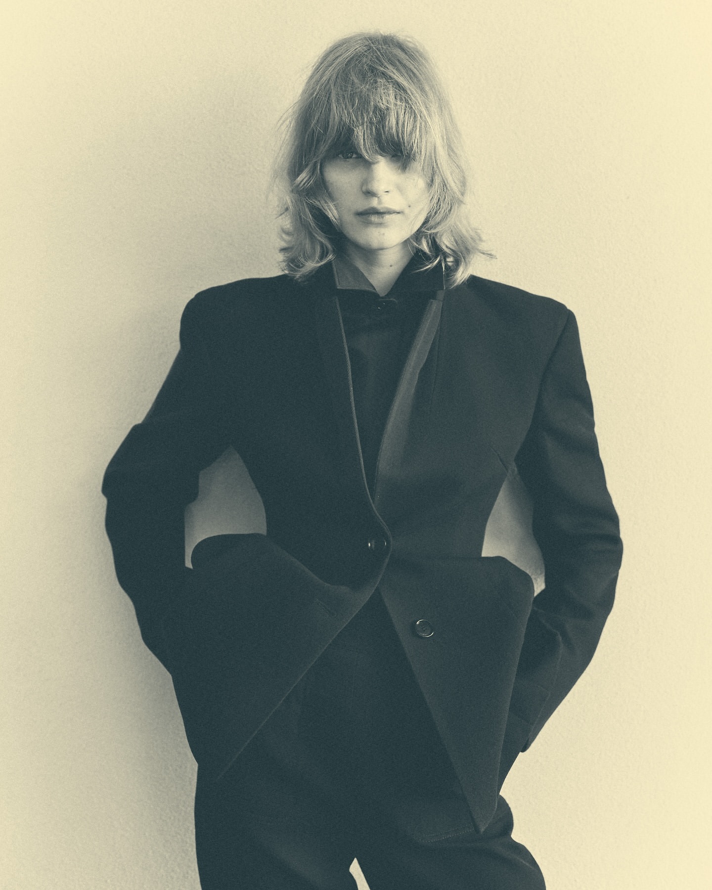 A stylish adult female model in a black suit stands in front of a simple off-white background, in a studio shot, showcasing fashion.