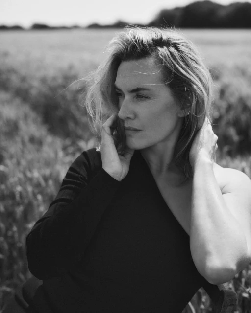 Black and white portrait of an adult woman with short hair sitting in an open field looking off to the side on a sunny day.
