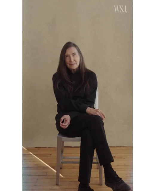 Portrait of woman in a black outfit sitting on a chair in front of a beige wall, lit by natural sunlight.