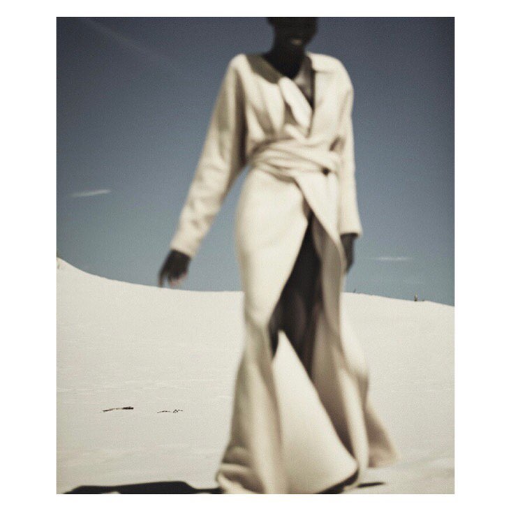Adult female in a long, wrap-style coat dress walks on sand dunes under a clear sky. The color palette is neutral with soft lighting.