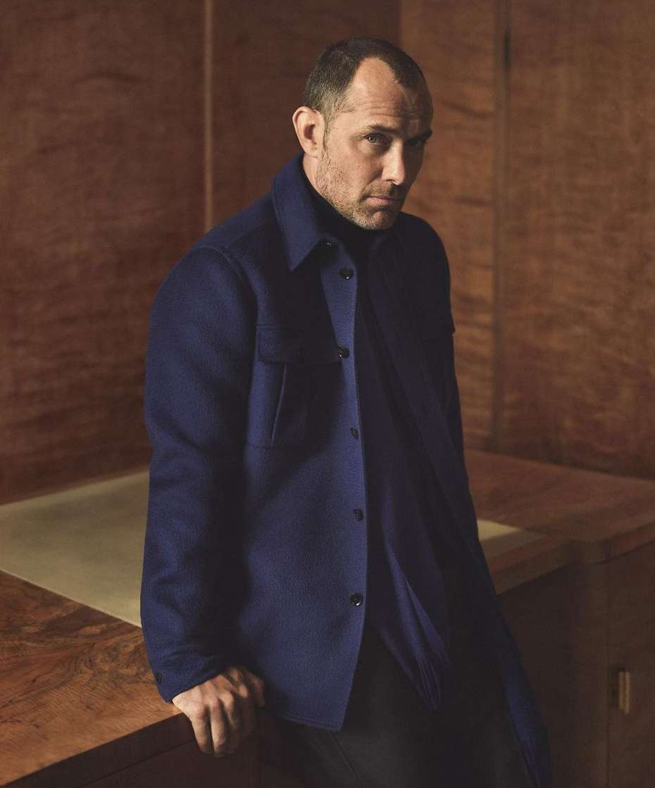 Portrait of Jude Law wearing a dark blue button-down shirt standing in a room with wooden walls.