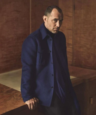Portrait of Jude Law wearing a dark blue button-down shirt standing in a room with wooden walls.