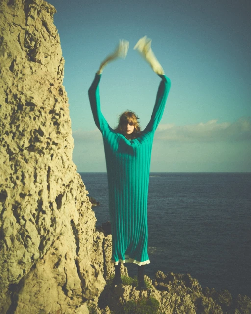 A slender adult fashion model with blonde hair poses on a coastal cliff in a long, teal, ribbed dress with her arms raised overhead.