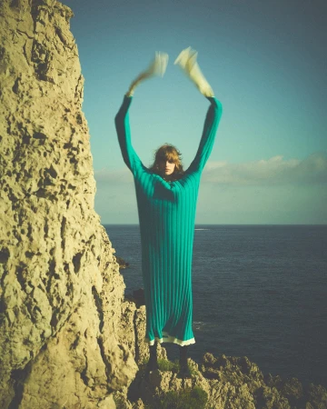 A slender adult fashion model with blonde hair poses on a coastal cliff in a long, teal, ribbed dress with her arms raised overhead.