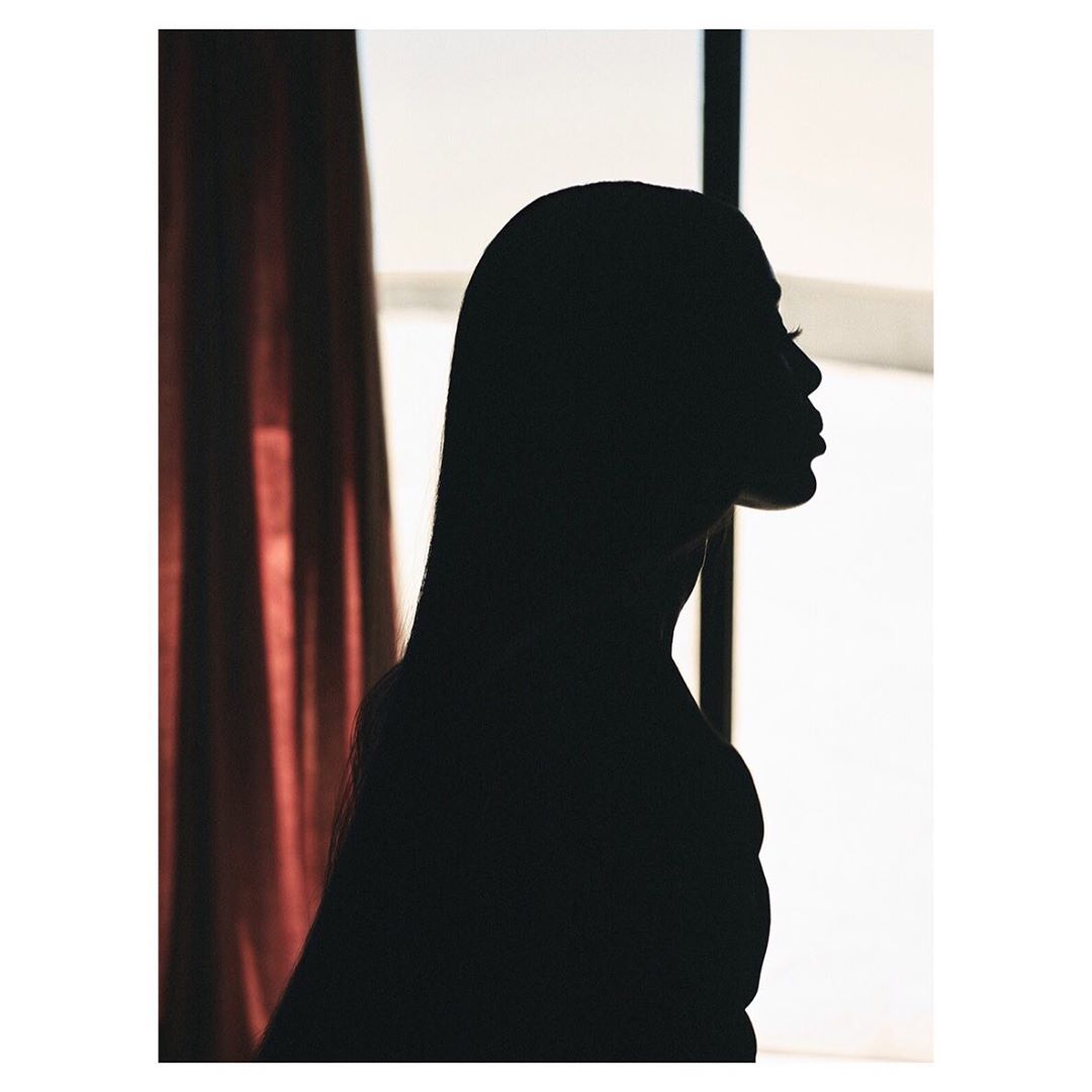 Silhouette of a woman with long hair in profile with a window and red curtain in the background, creating a dramatic chiaroscuro effect.