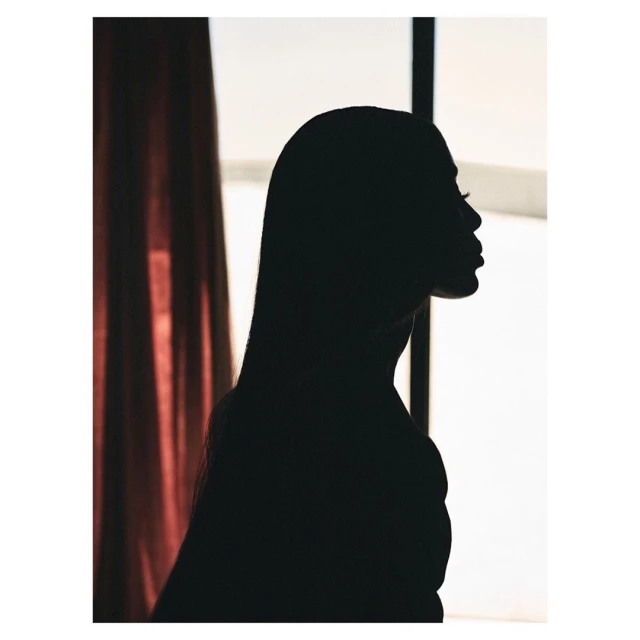Silhouette of a woman with long hair in profile with a window and red curtain in the background, creating a dramatic chiaroscuro effect.